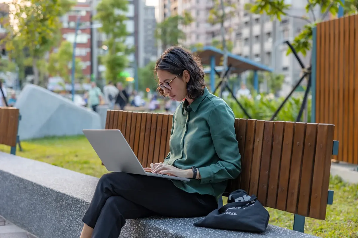 A woman using a laptop on a bench in a city park, embracing remote work outdoors.